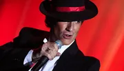 A performer in a black hat and suit is singing into a microphone against a red background.