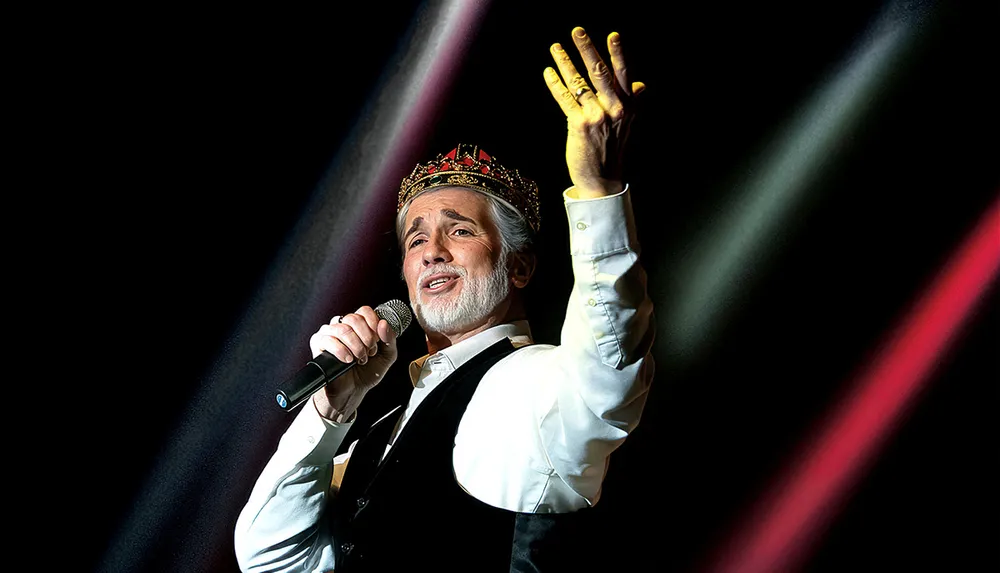 A person wearing a crown is passionately singing into a microphone on stage illuminated by colored spotlights