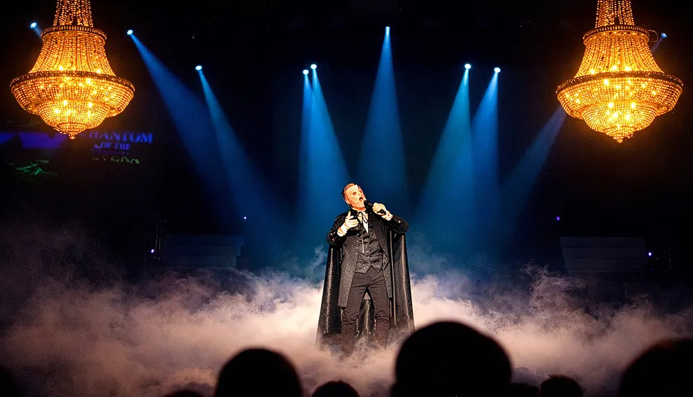 A performer wearing a mask and cape sings on a smoke-filled stage with dramatic lighting and two large chandeliers above