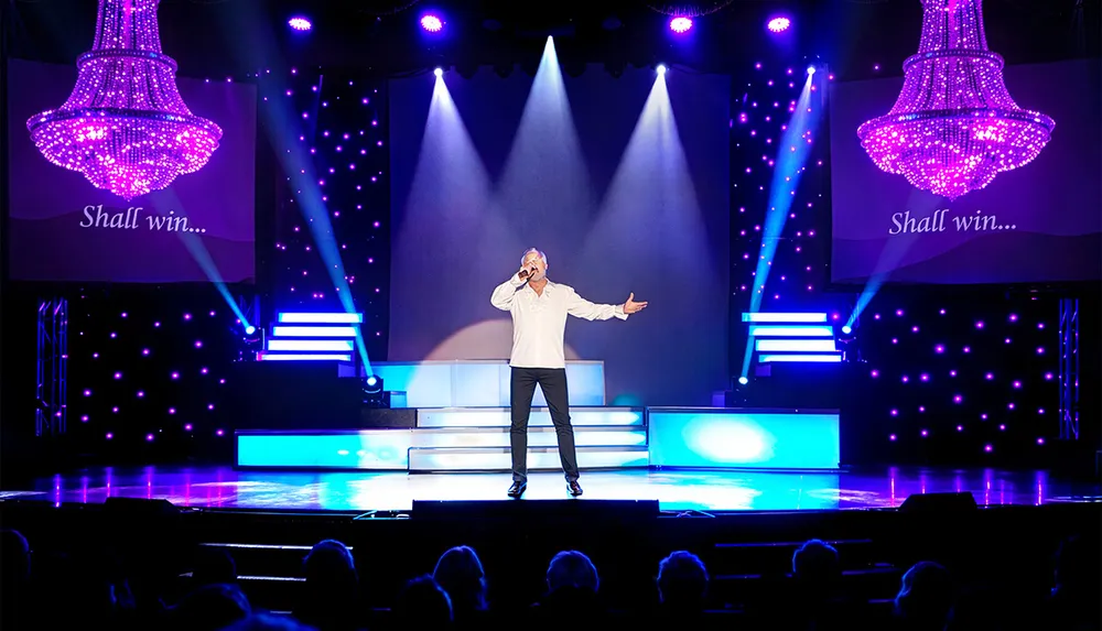 A performer is singing on a brightly lit stage with chandeliers and Shall win displayed on screens