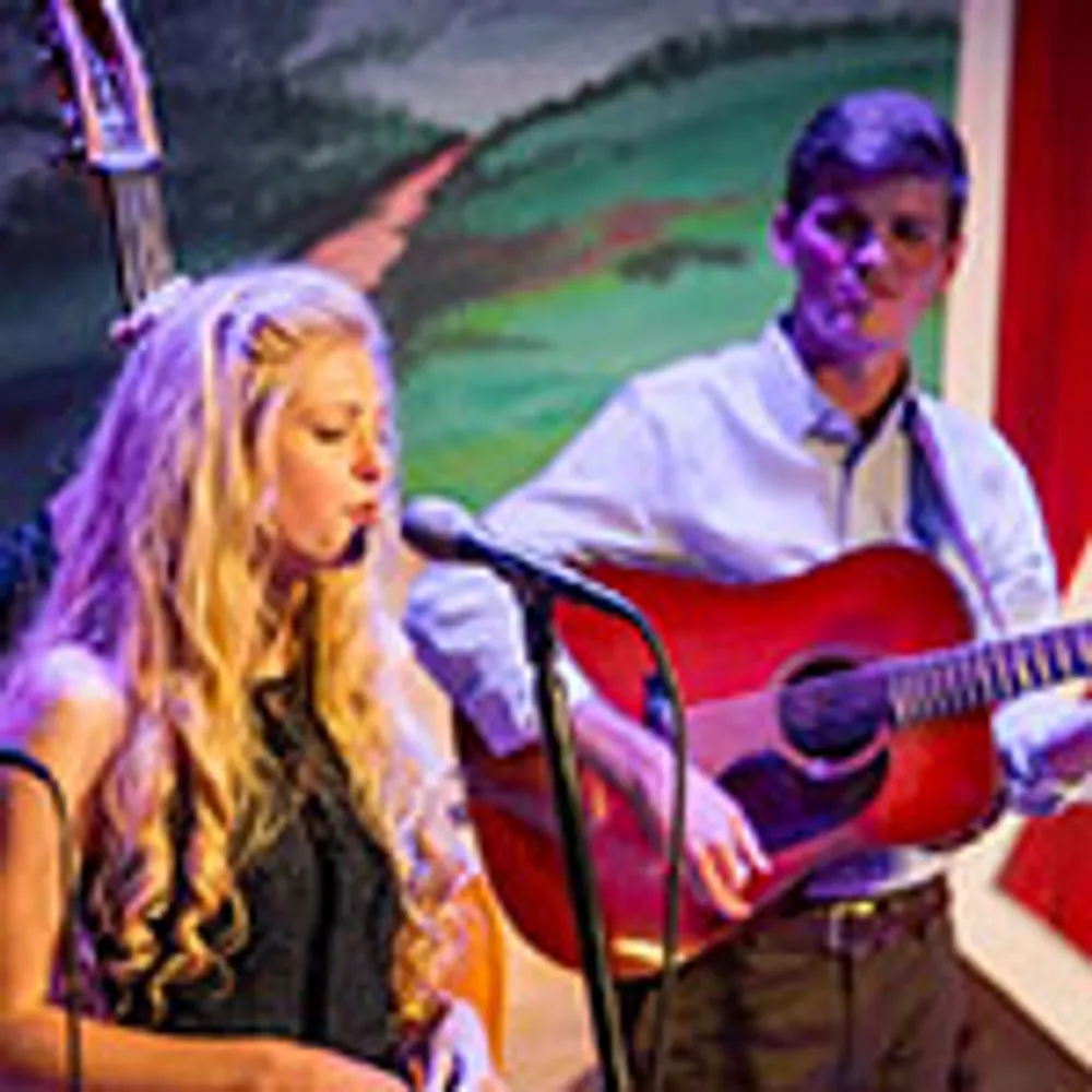 A woman sings into a microphone while a man plays the guitar on stage with colorful artwork in the background