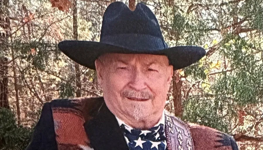 An elderly man with a mustache wearing a cowboy hat and a bandana with an American flag pattern smiles in front of a wooded background.