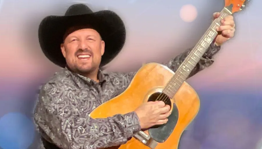 A smiling man wearing a cowboy hat plays an acoustic guitar against a softly blurred background.