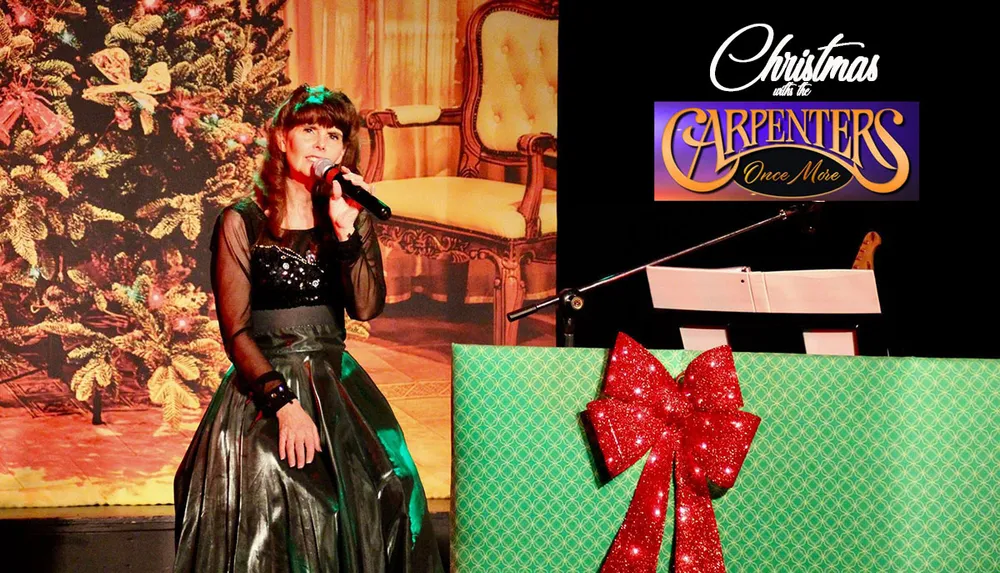 A person is performing on stage with a microphone in hand against a festively decorated backdrop with a Christmas tree and a large gift-wrapped box