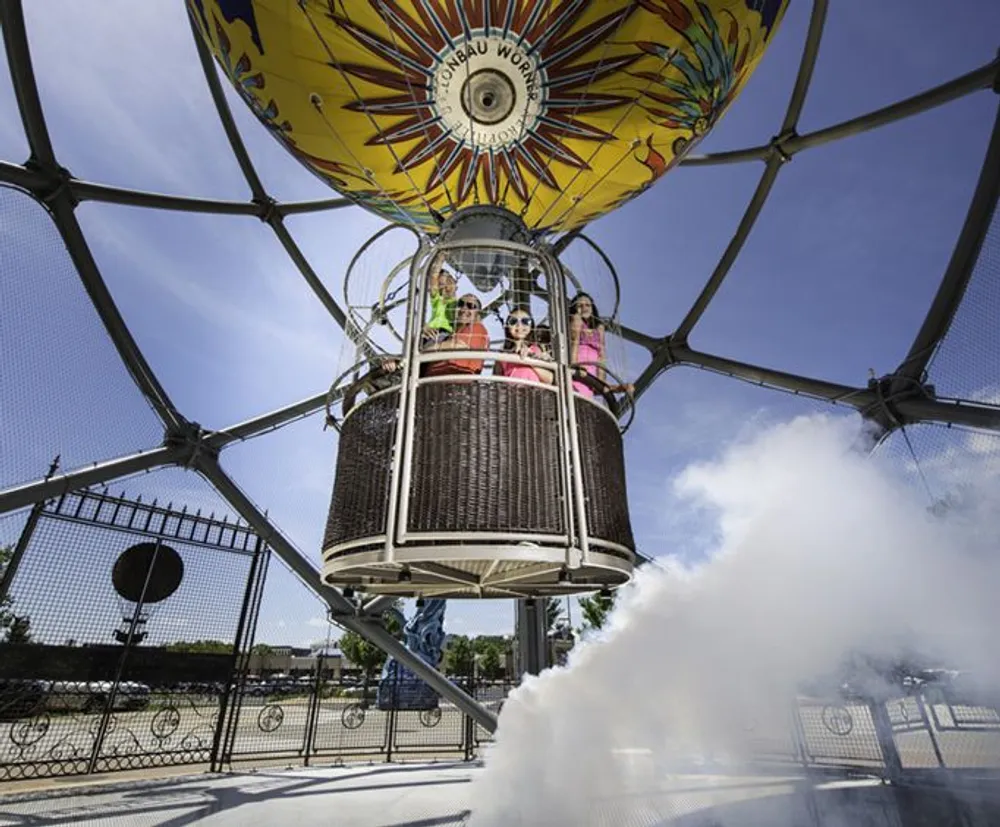 A group of people is riding in a hot air balloon basket secured under a colorful envelope as steam or smoke is released from a vent at the bottom left of the photo