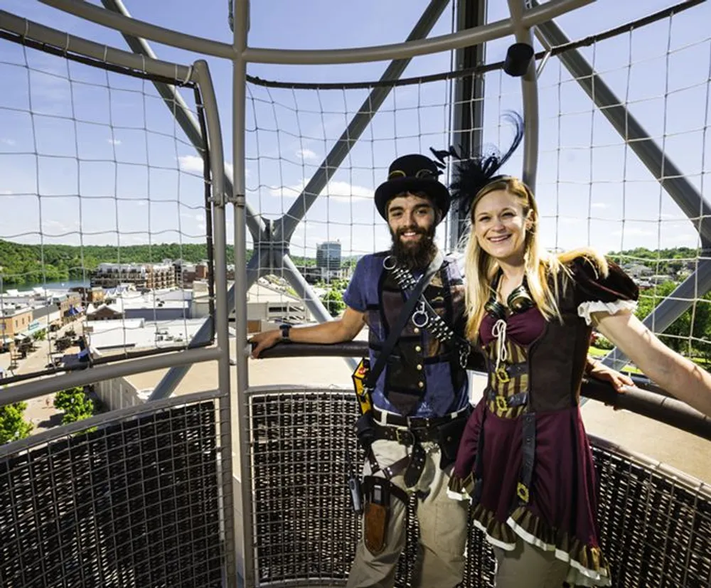 Two people in costume are smiling on an observation tower with a scenic urban background