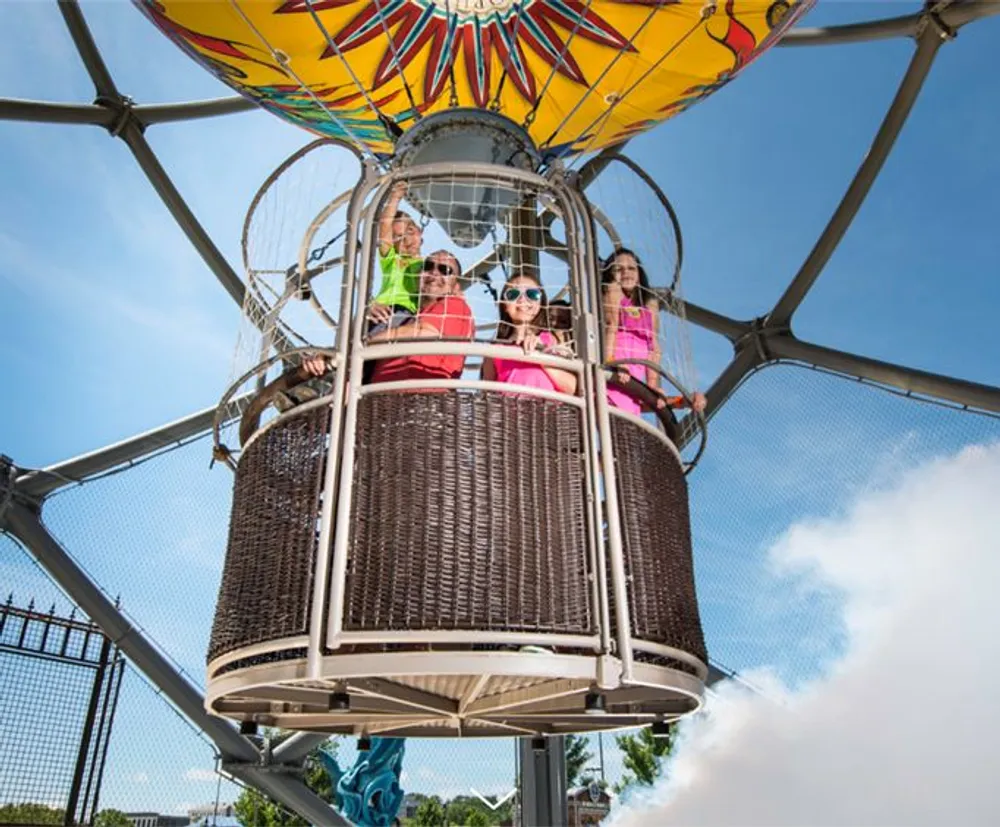 The image shows a group of people enjoying a ride in a colorful decorative hot air balloon basket at a theme park or fairground under a clear blue sky