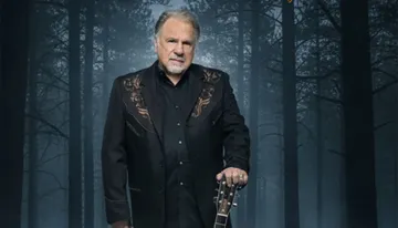 A man in a dark embroidered jacket stands in a misty forest setting, holding a guitar.