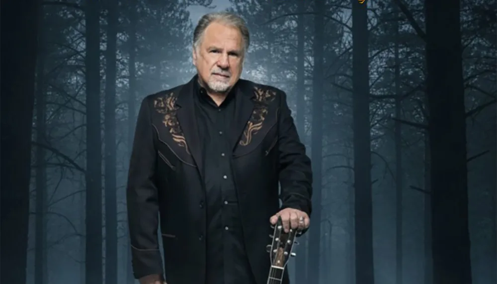 A man in a dark embroidered jacket stands in a misty forest setting holding a guitar