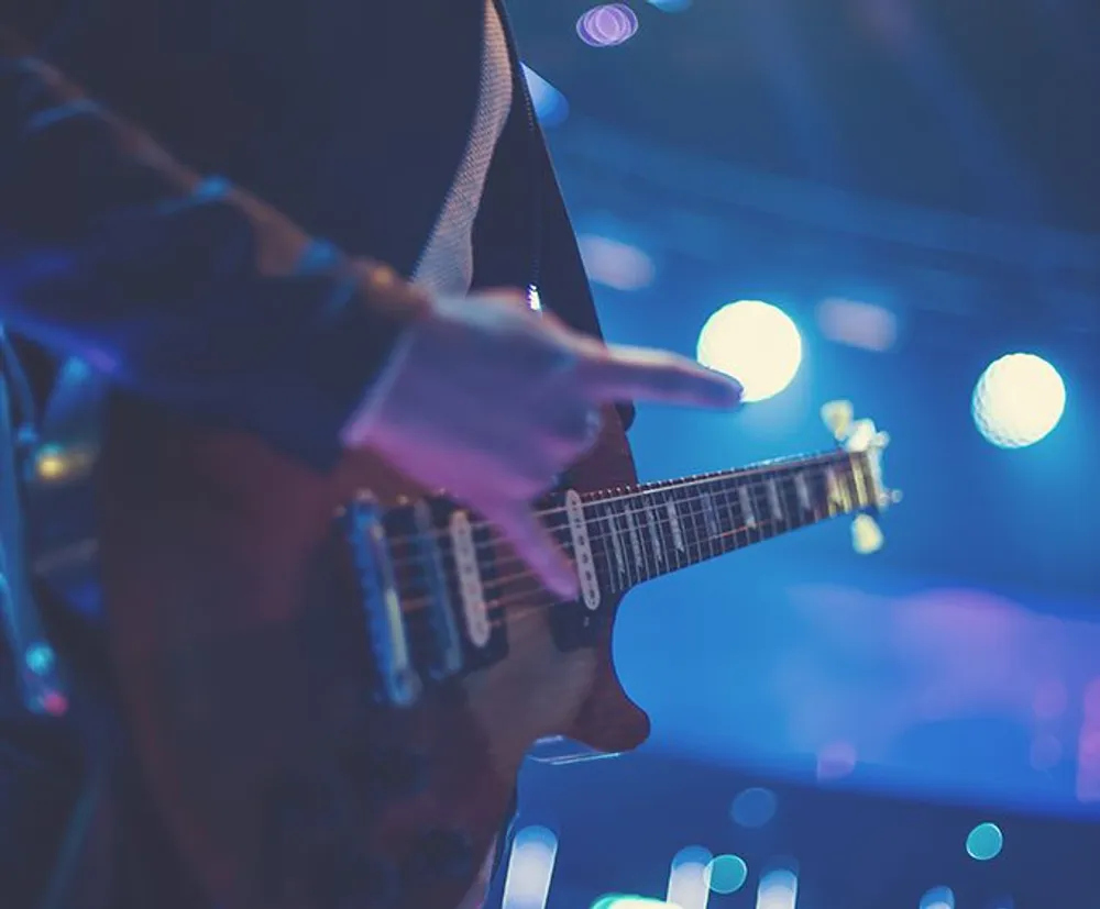 A guitarist is playing an electric guitar on stage with blurred lights in the background