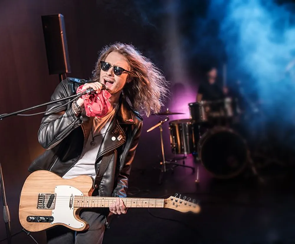 A rock musician holding a guitar passionately sings into a microphone on stage with blue stage smoke and a drummer in the background