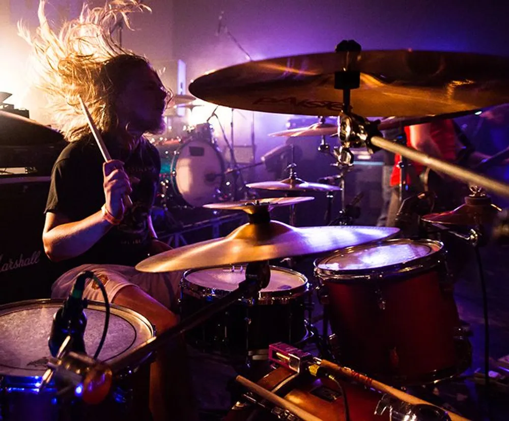 A drummer with flowing hair is passionately playing at a concert with a dramatic lighting setup highlighting the performance