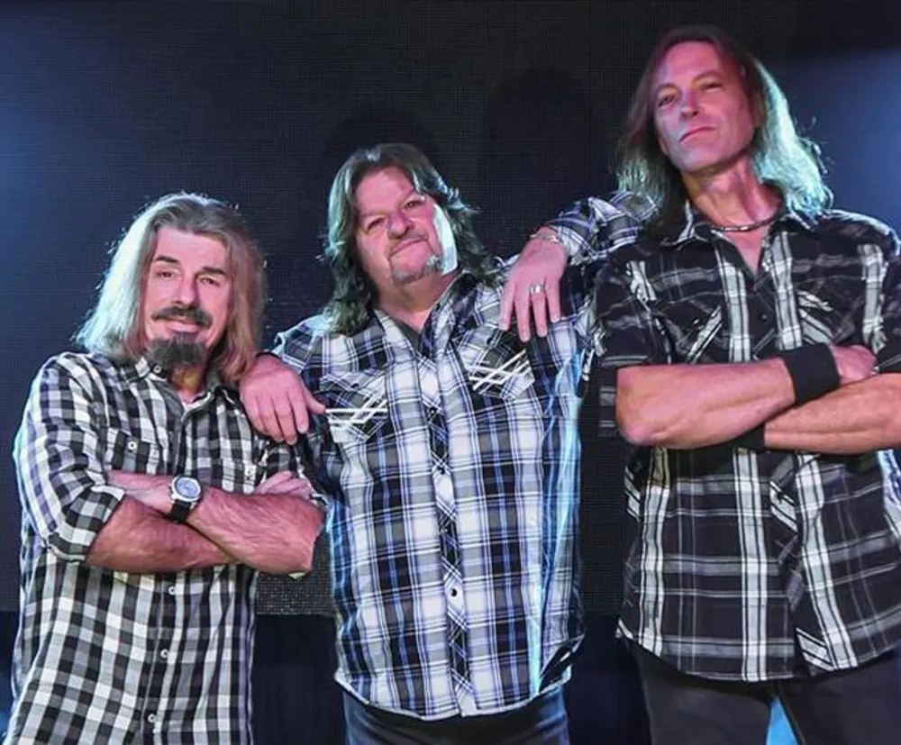 Three men with long hair wearing matching plaid shirts are posing confidently together on a stage with a blue-lit background