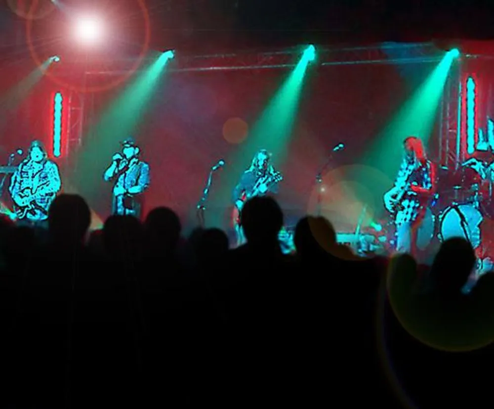 A band is performing on stage illuminated by colorful lights as silhouettes of an audience are seen in the foreground