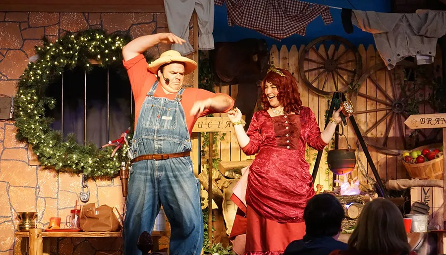 Two performers in festive costumes act on stage, surrounded by rustic decor, while an audience watches.