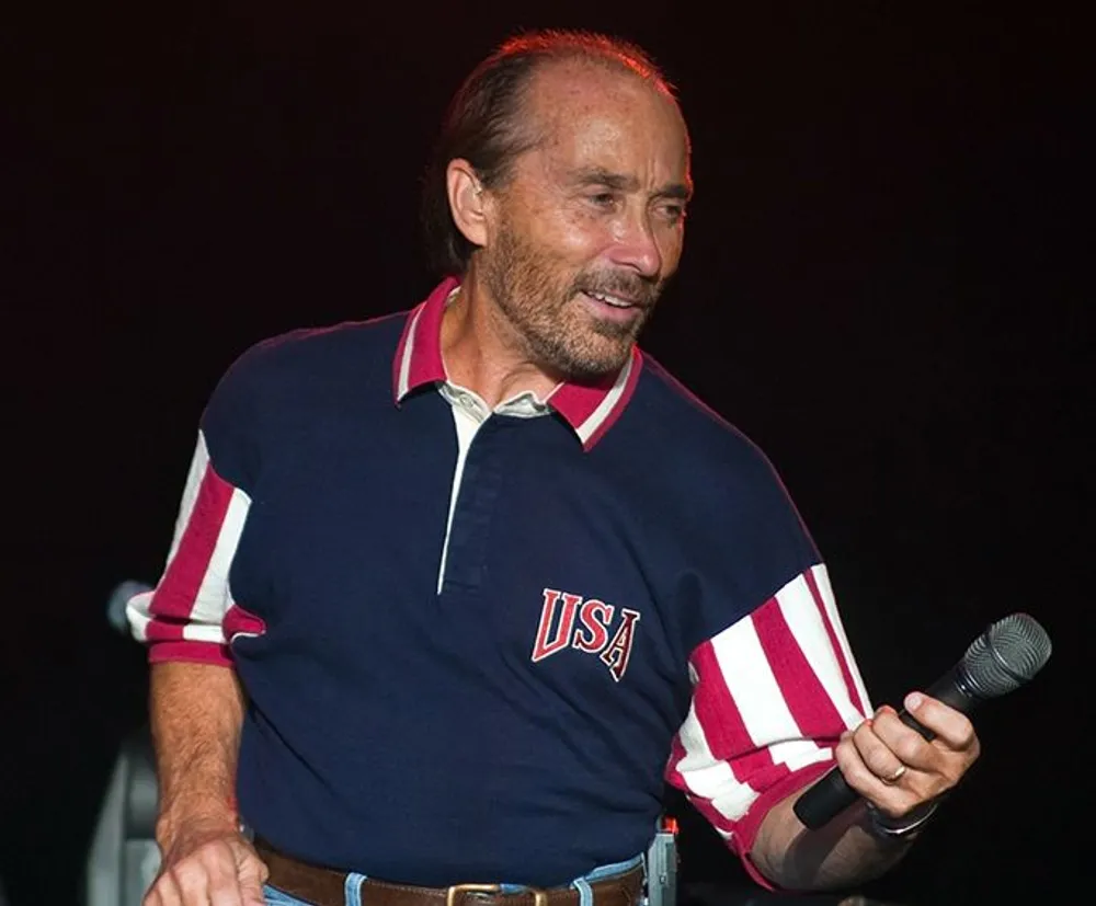 A man in a USA polo shirt is smiling and holding a microphone on a stage
