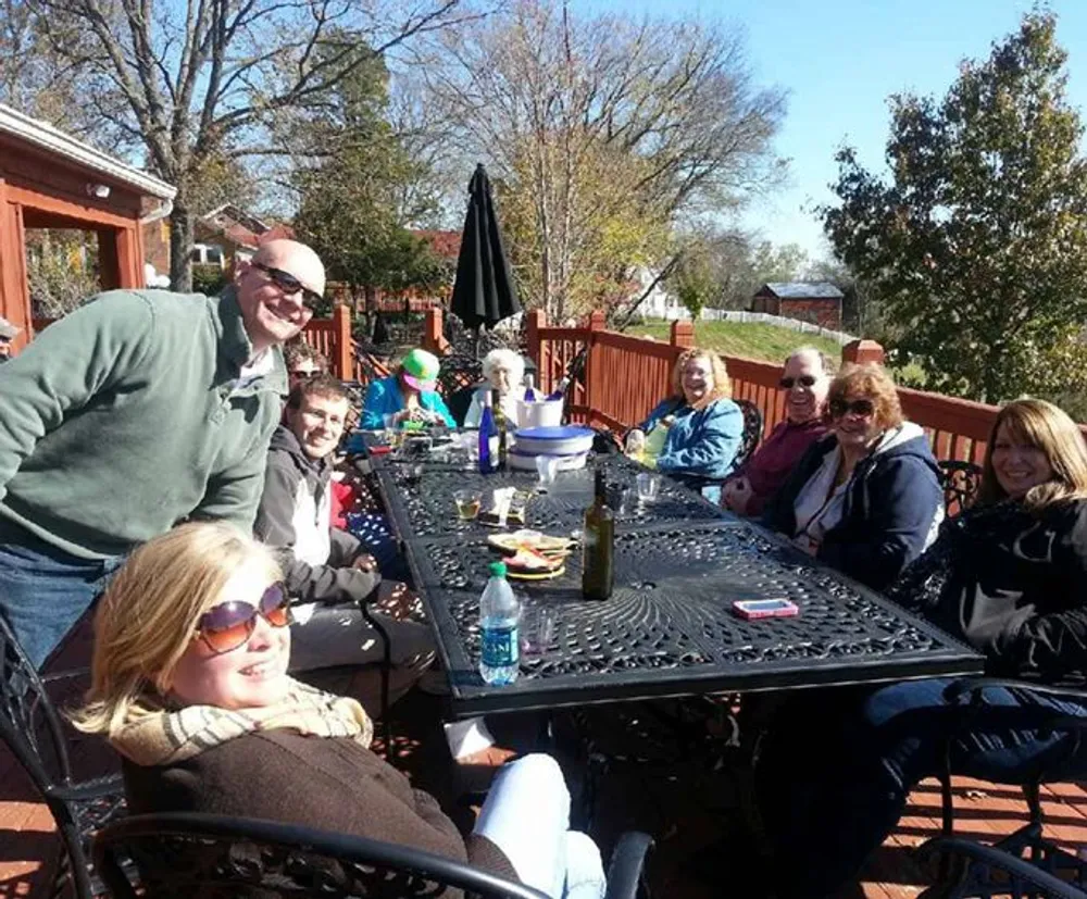 A group of people is sitting around an outdoor table smiling and enjoying what appears to be a sunny day gathering