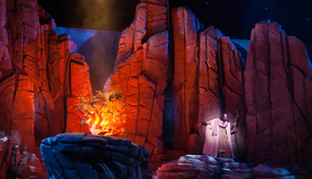 The image depicts a theatrical scene with a person in a white robe standing among stylized rocky structures with a burning bush effect to one side