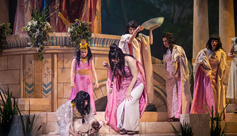 The image shows a theatrical performance with actors in ancient Egyptian-style costumes on a stage set designed to resemble an Egyptian temple