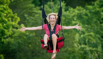 A person is joyfully zip-lining with arms outstretched against a backdrop of lush green trees.
