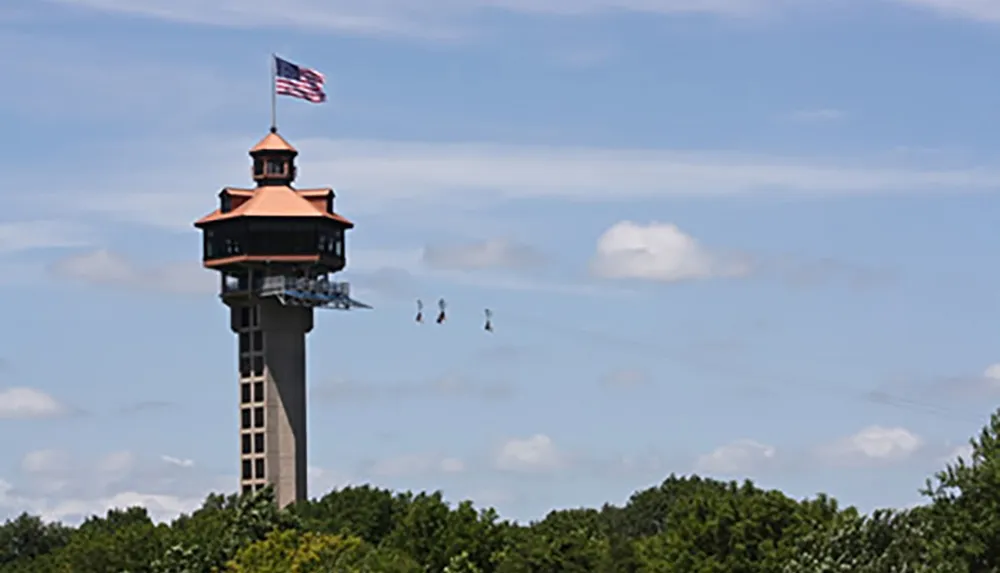 The image shows a tall observation tower with an American flag on top and a zip line extending from it with three people zip-lining above the treetops under a clear blue sky