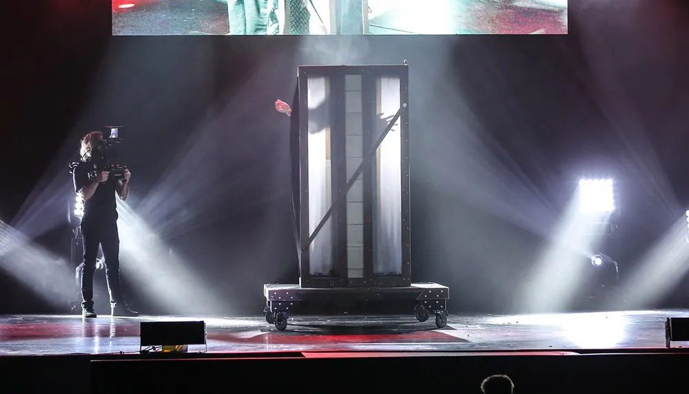 A camera operator is capturing a stage performance featuring a mysterious illuminated box amidst dramatic lighting