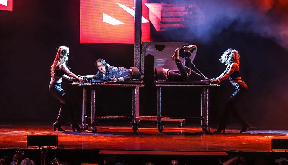 A magician lies on a table with the illusion of being cut in half while assistants interact with each part of the body against a dramatic stage backdrop with theatrical lighting and smoke effects