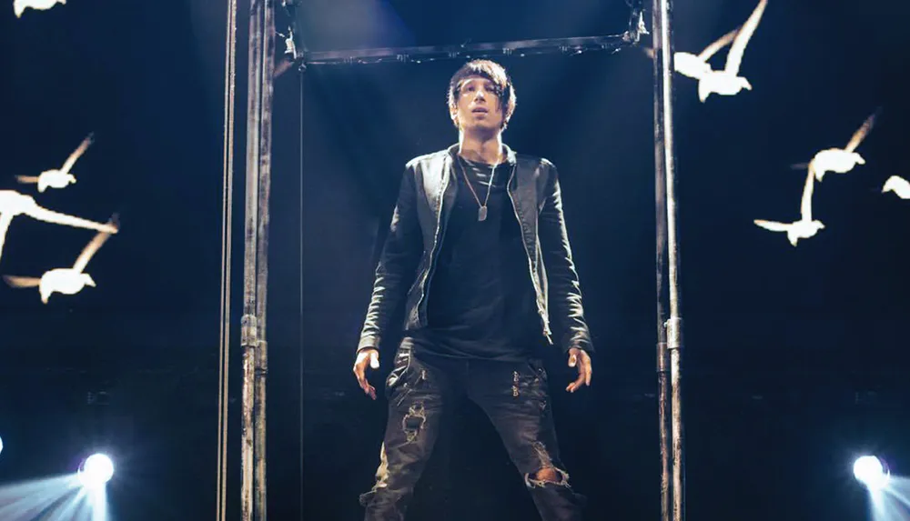 A person stands centered on a stage with dramatic lighting and a partially visible flying bird prop above creating an intense and theatrical atmosphere