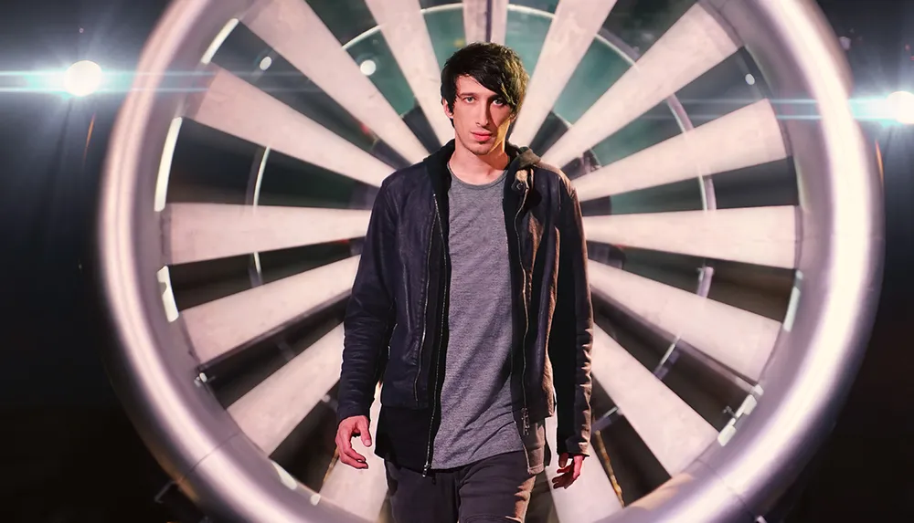 A young man stands confidently in front of an illuminated circular structure that resembles a dartboard with lights flaring behind him