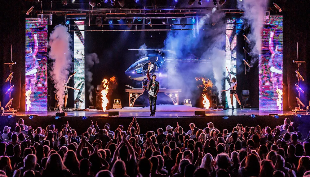 A performer stands on stage with dramatic lighting and effects including fire and a helicopter in front of a large audience