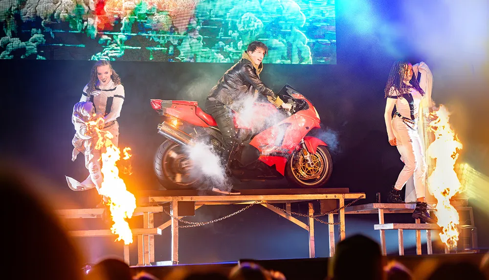 A performer on a red motorcycle is surrounded by flames and smoke on a stage with two other people in costume