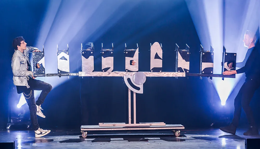 Two performers are on stage with a segmented platform seemingly performing a magic trick with one performer inside the compartments