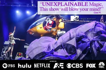 A performer stands on stage next to a helicopter with dramatic lighting, a flowing fabric, and a text quote praising an 
