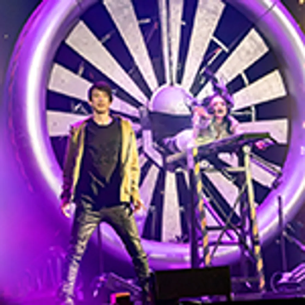 The image shows two performers on stage one is in front of a large fan-like structure under purple lighting