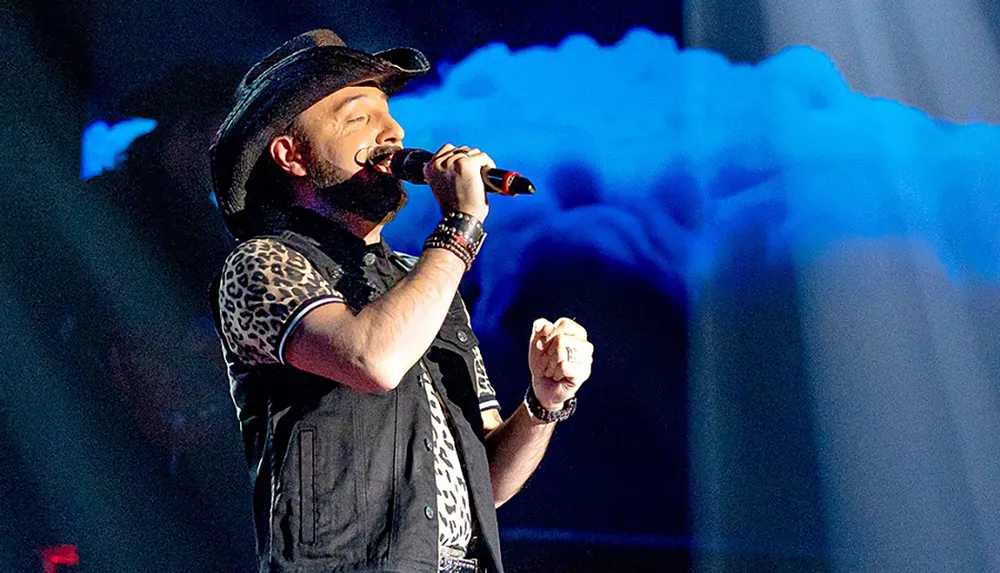 A man in a cowboy hat and leopard-print shirt passionately sings into a microphone on stage with dramatic blue lighting in the background