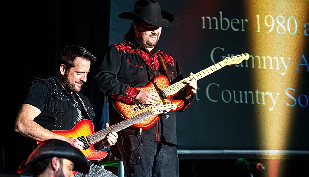 Two musicians are playing electric guitars on stage with one wearing a black cowboy hat and a black and red shirt