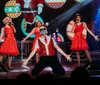 A group of performers in festive red outfits are energetically singing and dancing on stage with a Christmas-themed backdrop