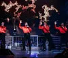A group of performers in festive red outfits are energetically singing and dancing on stage with a Christmas-themed backdrop
