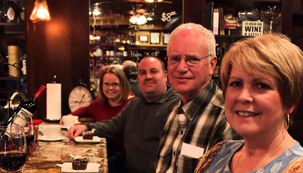 Four people are smiling at the camera in a cozy wine bar setting with a decanter of red wine and desserts on the counter in the foreground