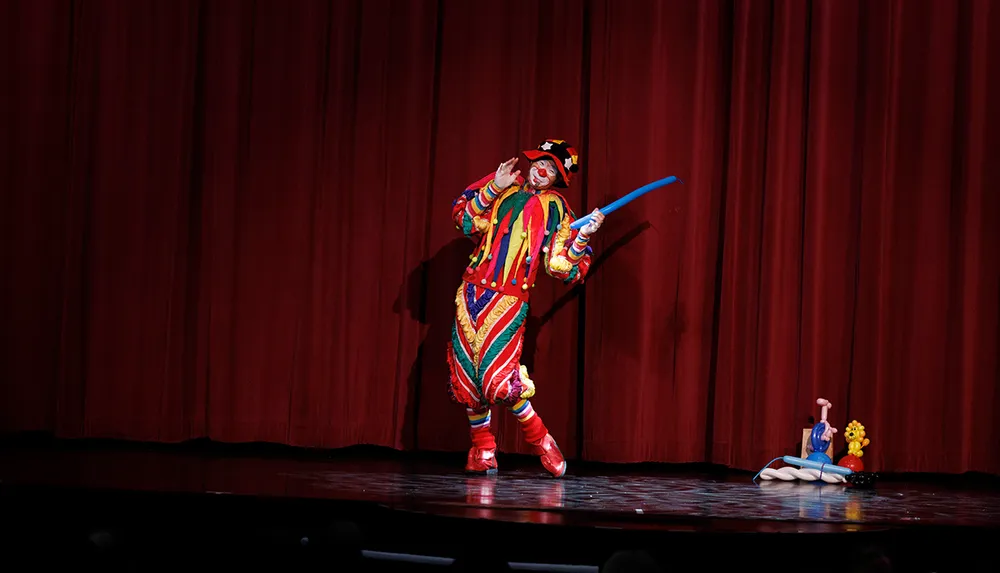 A performer in a colorful clown costume is on stage wielding a blue balloon sword with the audience in the foreground and a red curtain as the backdrop