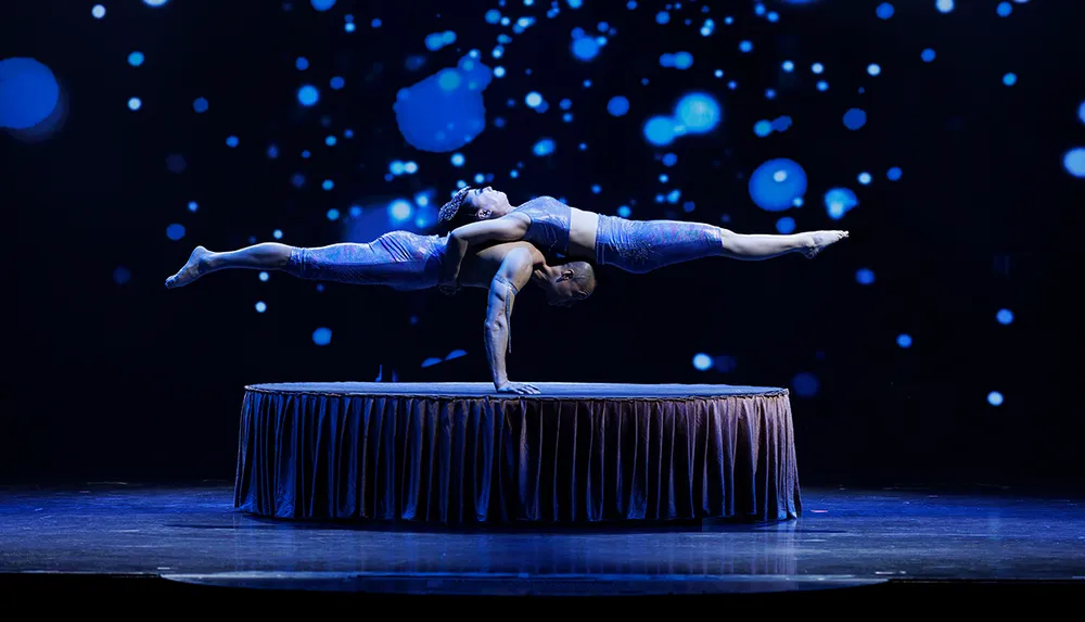 Two acrobats perform a dramatic balancing act on a circular platform under a mesmerizing array of blue lights