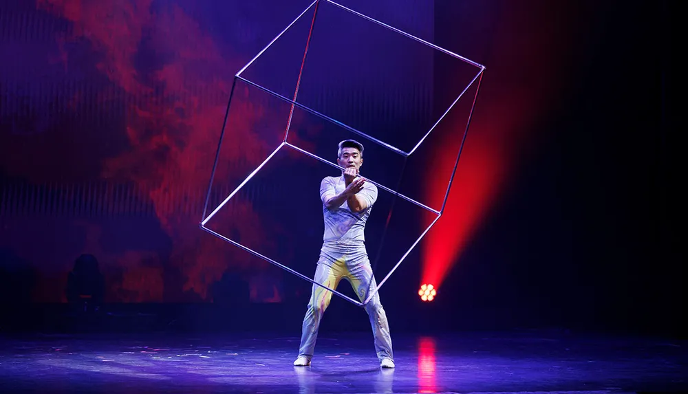 A performer on stage skillfully manipulates a large cube illuminated by a red spotlight