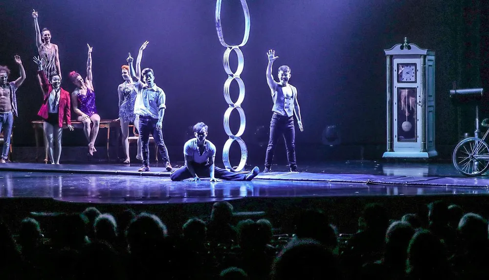 Performers are on a stage engaging in a theatrical or circus act with some raising their hands and one person in a split against a backdrop featuring a clock and bicycle parts
