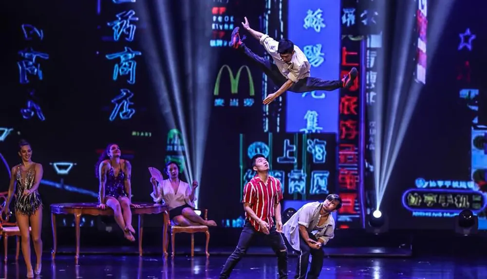 The image shows acrobats performing a dynamic routine on stage with one person mid-air above a group of others and a vibrant neon-lit urban backdrop