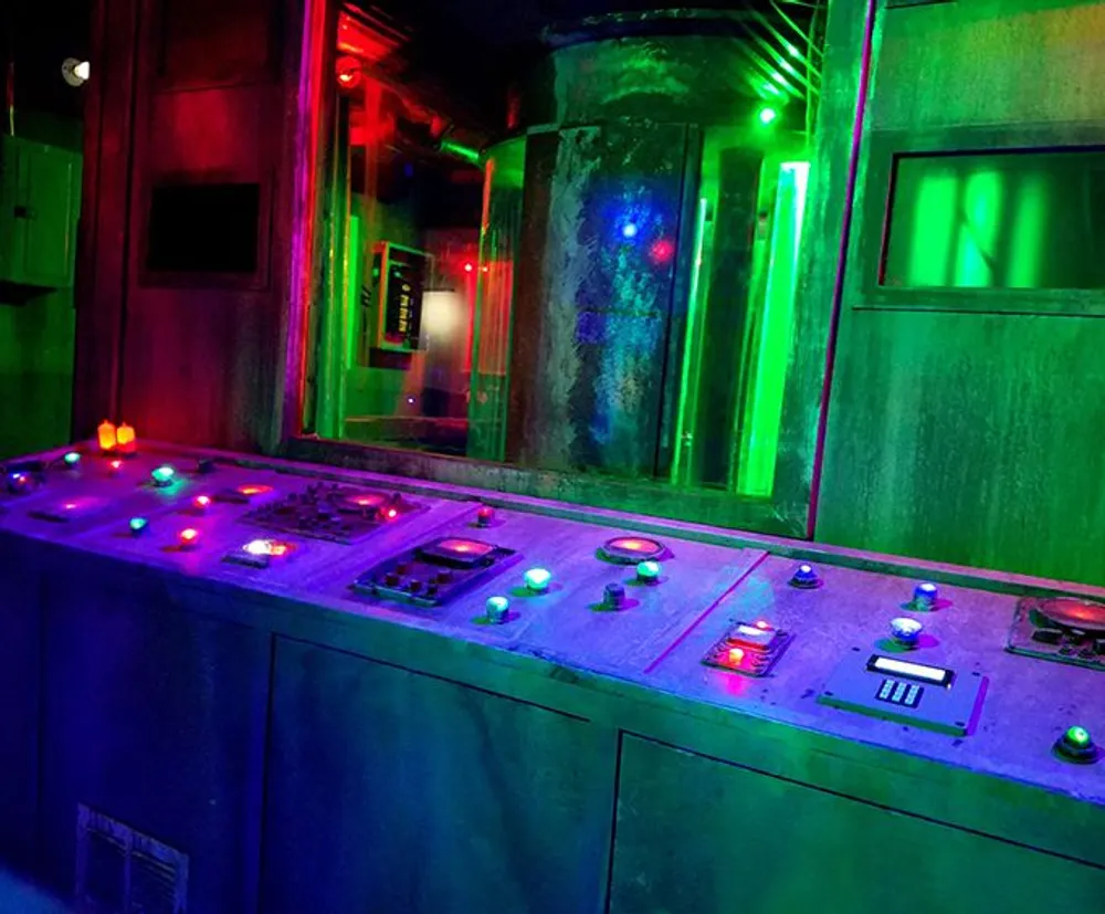 A control panel with various buttons and lights is illuminated by moody green and purple lighting giving it an industrial perhaps science fiction-like ambiance