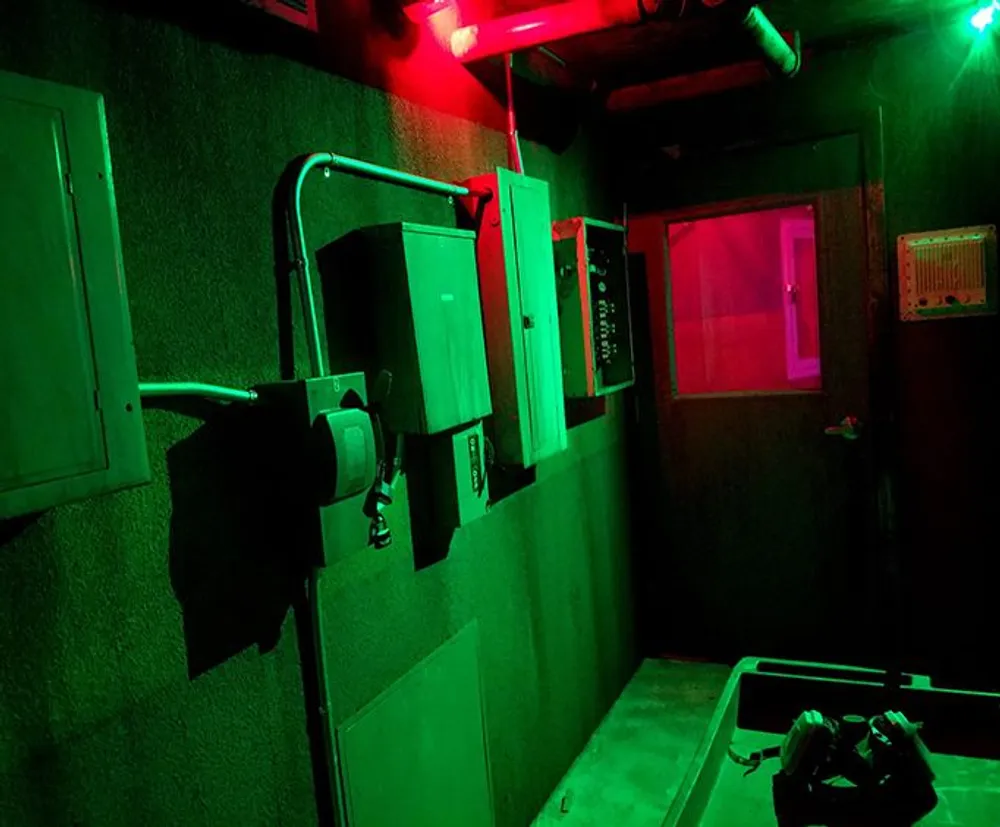 The image shows a dark room illuminated by red lights with several electrical panels and conduits mounted on the wall creating an industrial appearance