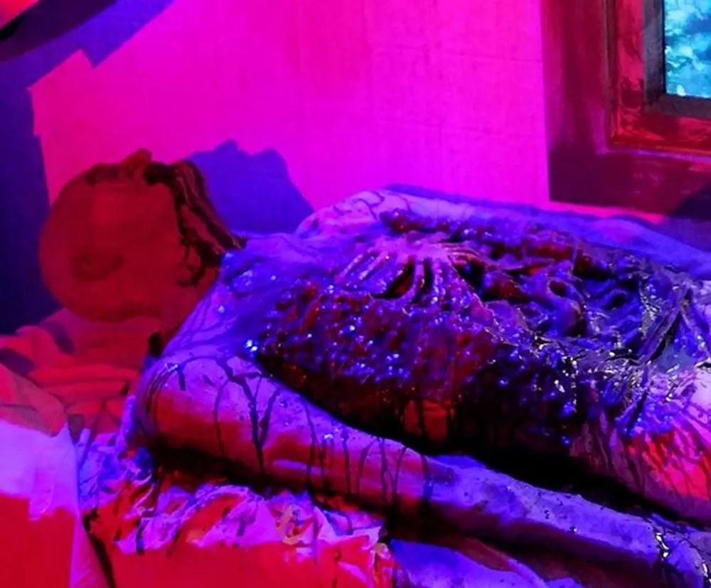 The image shows a horror-themed setting with a disturbing figure that looks like a decomposed body lying in a dramatic pose under purple and red lighting
