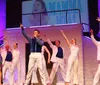 Five performers in white suits joyfully dance with top hats in hand on a stage with a purple geometric backdrop