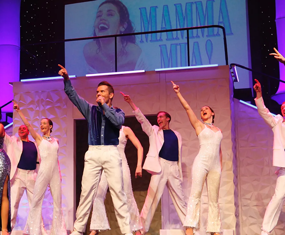 Performers are singing and dancing on stage during a production of Mamma Mia showcased by the backdrop and the attire of the cast