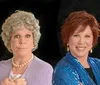 Two women one portraying an older character with gray hair glasses and a purple sweater and the other with reddish hair and a blue sequined jacket are side by side against a black background